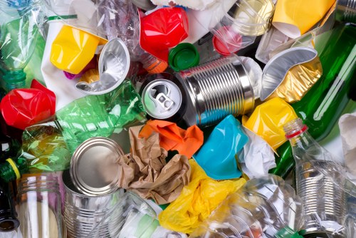 A close-up view of a large pile of mixed waste materials, predominantly consisting of crushed and crumpled plastic bottles in various colors including green, clear, red, yellow, and blue, some with caps still attached. There are also several metal cans, some with open and flattened tops, along with pieces of brown paper and cardboard among the plastics. The waste is spread across a surface that suggests an outdoor or industrial setting, with no clear background visible. This image reflects typical rubbish encountered in commercial waste removal services, such as those offered by Commercial Waste Mitcham, which operates in areas including Mitcham and nearby postcode regions. The collection of different materials highlights the importance of proper disposal and recycling processes for rubbish removal companies focusing on waste management in the local community.