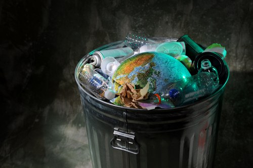 A close-up of an open metallic rubbish bin filled with various waste items, including plastic bottles, aluminium cans, and crumpled paper, with a small globe figurine placed among the debris. The bin is situated against a dark, textured background, and the waste materials vary in colour from transparent and green plastics to silver and brown paper. The scene highlights general household or office waste, with the globe serving as a symbolic element. The metallic surface of the bin has a reflective finish, and the image emphasizes waste collection relevant to rubbish removal services such as those offered by Commercial Waste Mitcham in the Mitcham area. The overall atmosphere is utilitarian, focusing on waste management and environmental responsibility.