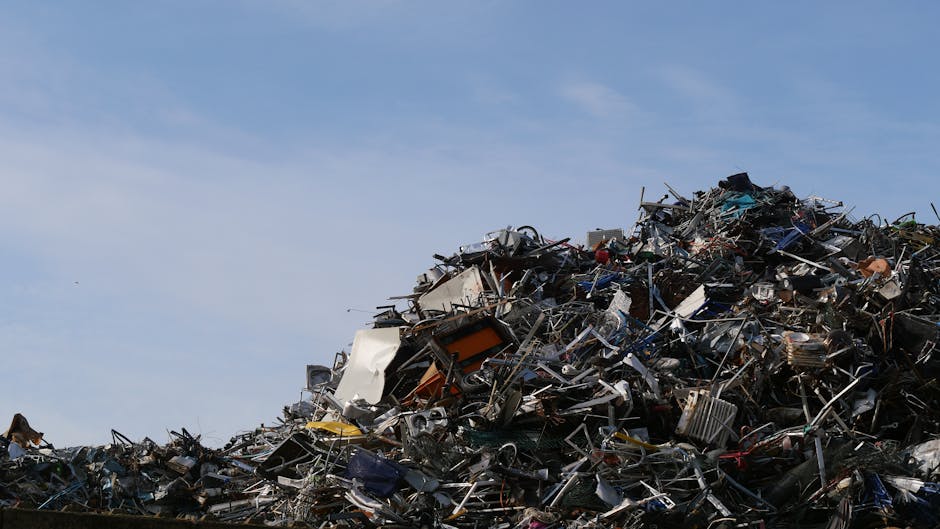 A large heap of assorted metal and electronic waste piled high outdoors against a clear blue sky. The jumble includes broken appliances, metallic scraps, wires, and components of various shapes and si